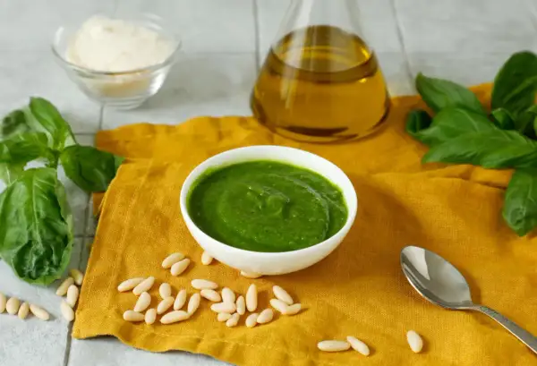 Pesto fresco in una ciotola bianca, con ingredienti per una ricetta pesto semplice: basilico, pinoli, olio d'oliva e parmigiano grattugiato.