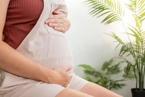 Ciężarna kobieta trzyma dłonie na brzuchu. W tle zielone liście roślin. Temat: rozwarcie szyjki macicy.