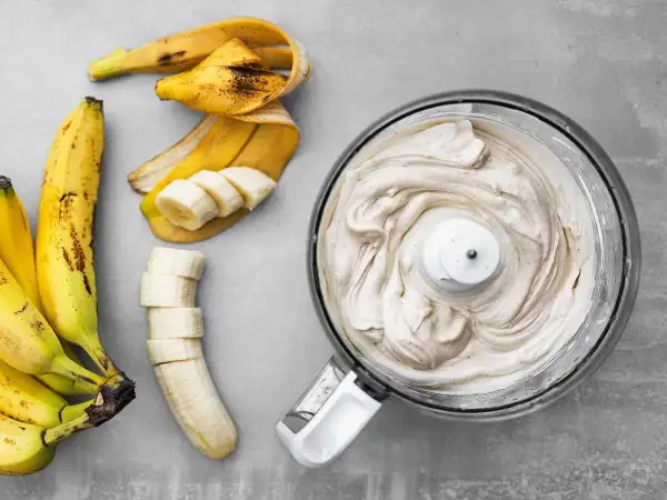 En matberedare fylld med krämig bananfrukt-glass bredvid skalade och skivade bananer.