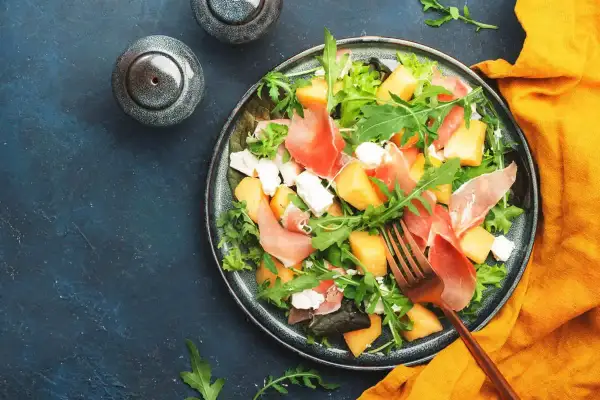 Insalata con melone, prosciutto crudo e formaggio fresco, guarnita con rucola. Un piatto estivo perfetto.