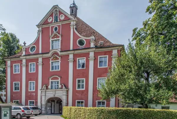 Prachtvolles Gebäude des Klosters Salem mit roter Fassade, weißen Säulen und einem geschwungenen Giebel. Ein Baum spendet Schatten.