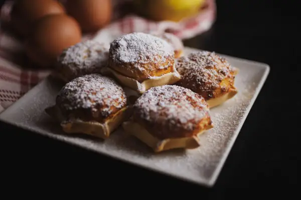 Cinque dolci sardi, spolverati di zucchero a velo, su un piatto bianco. Sullo sfondo, uova e un tessuto a quadri rossi.
