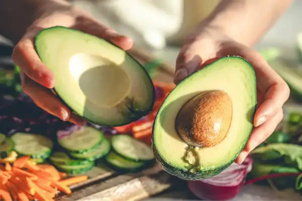 Manos sostienen un aguacate cortado por la mitad, listo para disfrutar. Es la temporada del aguacate en España, con verduras frescas de fondo.