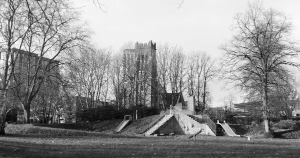 A playground with slides and stairs, captured on Ilford Pan 400 film, stands amidst bare trees with a brick tower in the background.