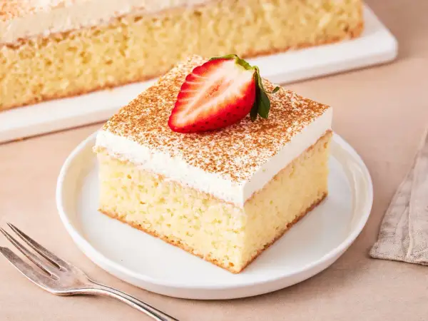 Ein Stück saftiger Tres Leches Kuchen, garniert mit einer Erdbeerhälfte und Zimt, auf einem Teller mit einer Gabel daneben.