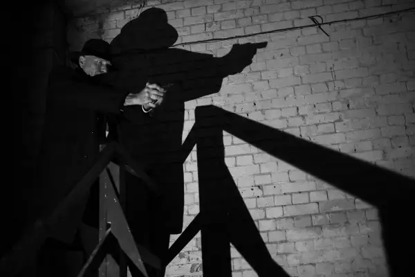 A man in a fedora and trench coat aims a pistol in this dramatic film noir photography. His shadow looms large against a brick wall.