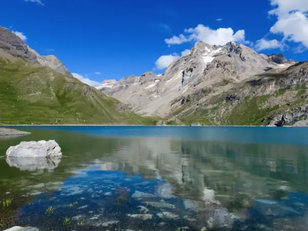 Le lac de la Sassière, aux eaux turquoise, reflète les montagnes majestueuses et le ciel bleu. Un rocher émerge, témoin de la quiétude du paysage.