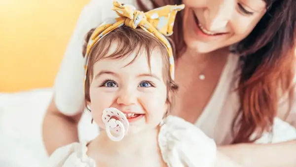 A baby with bright blue eyes and a yellow headband smiles, her passy teeth peeking out from behind a pacifier. Her mother smiles in the background.