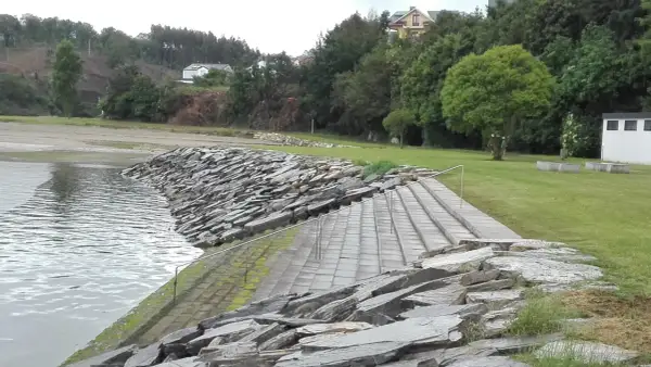 Escaleras de piedra y césped verde junto al agua en la playa de los bloques Ribadeo, con casas y árboles al fondo.