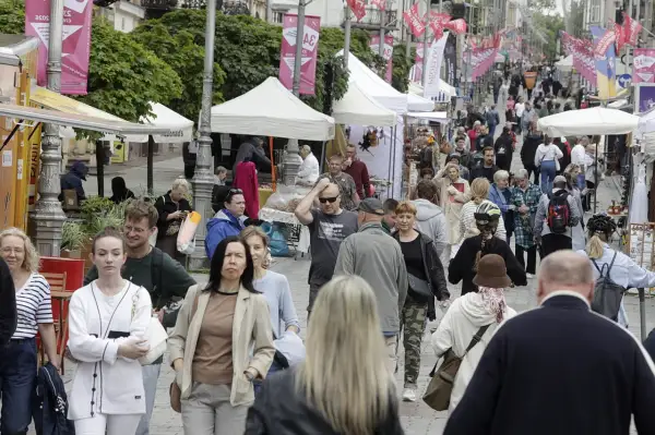 Tłum ludzi spaceruje ulicą z kramami i namiotami, ozdobioną flagami.