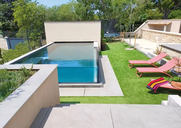 Un magnifique bassin vitré aux eaux bleues, entouré d'une pelouse synthétique et de chaises longues roses.