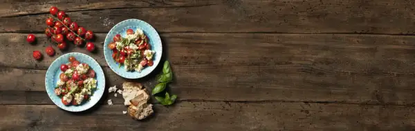 Två tallrikar med tomatsallad och vispad mozzarella, garnerad med basilika. Körsbärstomater och bröd på träbord.