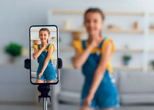 Dziewczynka na ekranie telefonu uśmiecha się, podczas gdy mama w tle nagrywa filmik. To przykład zjawiska sharenting co to znaczy dzielenie się życiem dzieci w sieci.