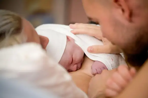 Pierwsze chwile po narodzinach. Noworodek otulony kocykiem, rodzice z czułością dotykają maleńkiego ciałka. Tak wygląda poród.