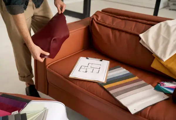 A person is choosing couch materials, holding a burgundy swatch next to a brown leather couch. Fabric samples and a floor plan are on the seat.
