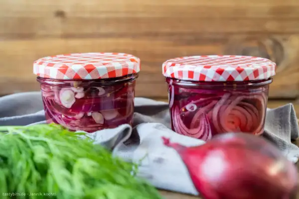 Zwei Gläser mit eingelegten roten Zwiebeln, bereit für Salate oder Sandwiches. Frischer Dill liegt daneben.