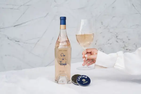 Une main tient un verre de vin rosé. À côté, une bouteille de Clos du Temple et une boîte de caviar. Une belle conservation rosé pour un moment d'exception.