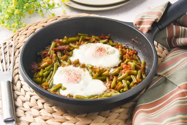 Judías verdes salteadas con trocitos de bacon y dos huevos fritos encima, servido en sartén.