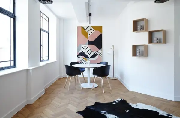 Un comedor moderno con mesa redonda, sillas negras y una alfombra geométrica. Las estanterías flotantes y las ventanas grandes dan la sensación de casas pequeñas y acogedoras.