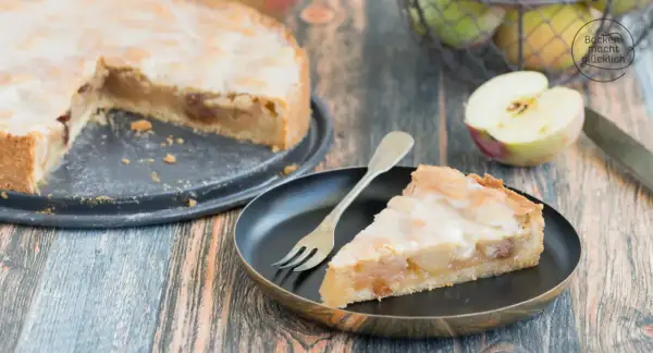 Ein Stück saftiger Apfelkuchen mit süßer Glasur und Mandelsplittern, daneben ein halber Apfel und ein Korb voller Äpfel.