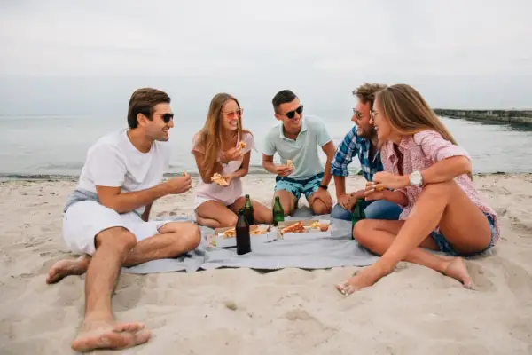 Grupa przyjaciół cieszy się piknikiem na plaży, jedząc pizzę i pijąc napoje. To świetny pomysł na wakacje!