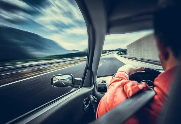 Kierowca w pomarańczowej kurtce jedzie autostradą. Widać rozmycie drogi i krajobrazu, co sugeruje dużą prędkość. Wymaga to ostrożności, a nawet szczególnej ostrożności.