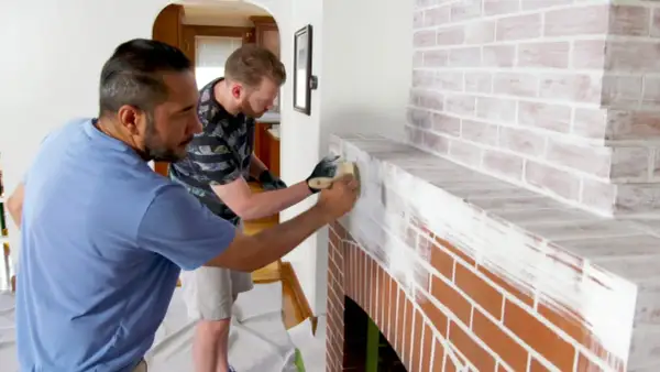 Two men are learning how to whitewash brick on a fireplace, applying white paint with brushes for a rustic look.