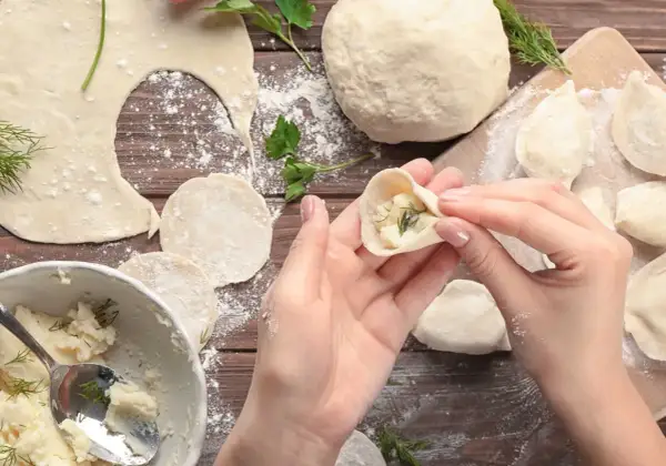 Ręce formują pieroga z ciasta na pierogi, wypełnionego farszem ziemniaczanym i koperkiem. Na drewnianym blacie widać mąkę, gotowe pierogi i miskę z farszem.