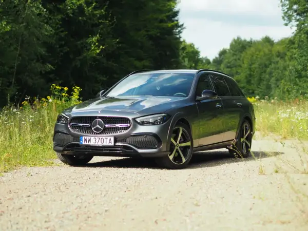 Szary Mercedes-Benz Klasy C All-Terrain na szutrowej drodze, otoczony zielenią lasu i polnymi kwiatami.