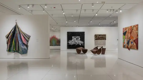 An art gallery features diverse works, including a colorful draped sculpture, a black and white textured piece, and a vibrant collage, reminiscent of the bold style of Pamela Joyner.