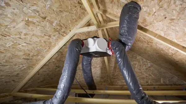 Installation d'une VMC dans un grenier. Le caisson blanc est suspendu, relié à des gaines grises flexibles.
