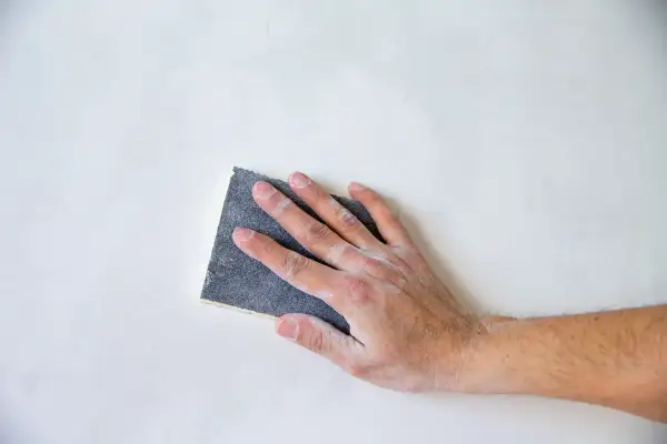 Mano che carteggia una parete bianca, un gesto che illustra il significato di carteggiare per levigare e preparare le superfici.