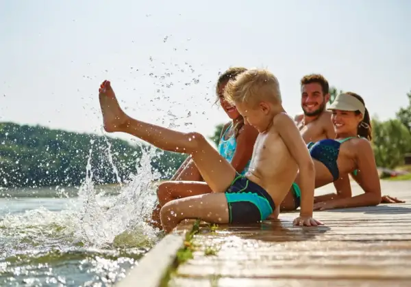 Familie genießt den Sommer am See. Kinder planschen im Wasser, Eltern entspannen sich. Guter uva schutz ist wichtig.