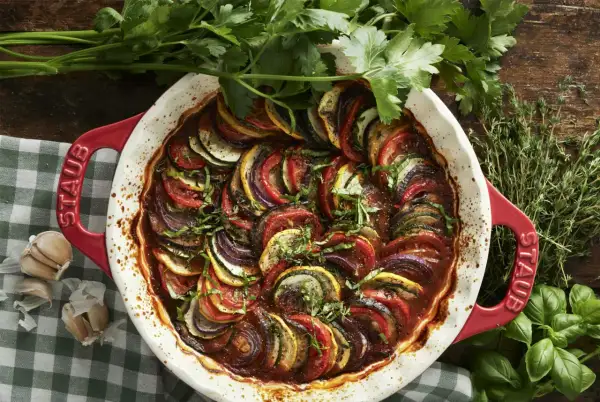 Vacker ratatouille i en röd ugnsform, med skivade grönsaker som tomat, zucchini och aubergine. Perfekt för ett ratatouille recept.