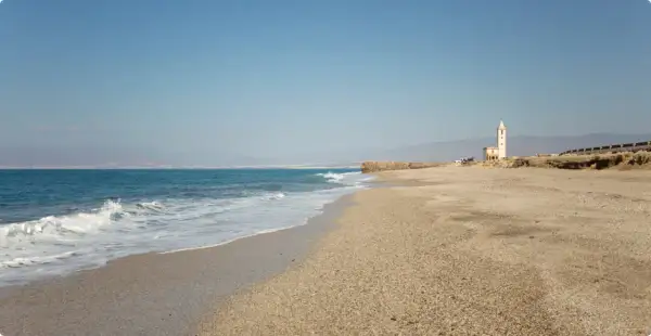 La almadraba de Monteleva, una torre blanca se alza en la playa, con el mar azul y las montañas al fondo.