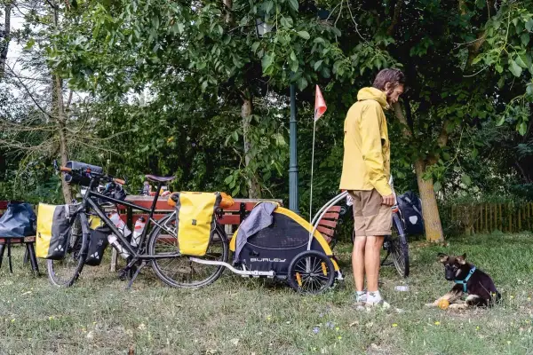 Un homme en jaune se tient près d'un vélo chargé, avec une remorque et un jeune chien joueur. Un chien Canada prêt pour l'aventure.