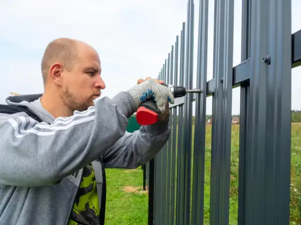 Mężczyzna w rękawiczkach przykręca wkrętarką element do nowego, szarego płotu.