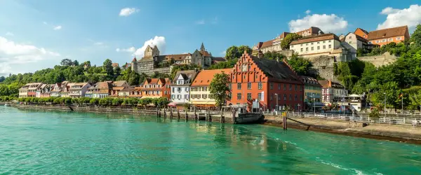 Meersburg Tourismus: malerische Altstadt mit bunten Häusern und Burg am Hang, überragt von grünen Hügeln, am glitzernden Bodensee.
