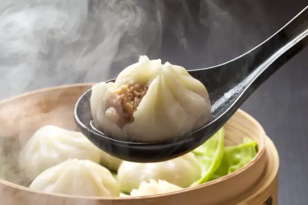 Gorące pierożki dim sum unoszą się w parze, jeden z nich na łyżce odsłania soczyste nadzienie.