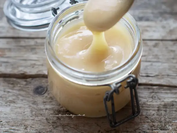 Un cucchiaio preleva il cremoso latte condensato da un barattolo di vetro su un tavolo di legno rustico.