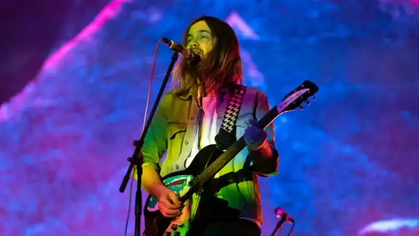Kevin Parker z Tame Impala gra na gitarze podczas koncertu, w tle kolorowe światła.