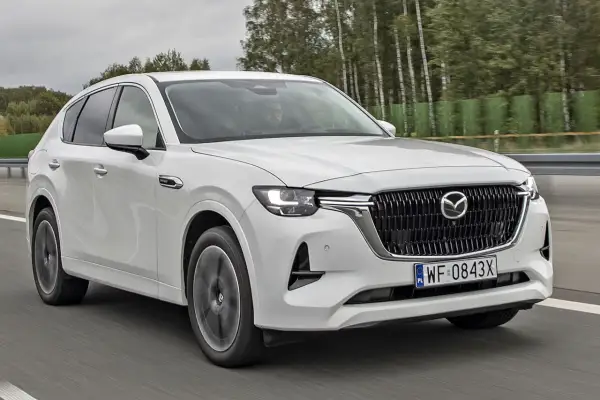 Biała Mazda CX-60 jedzie po drodze, otoczona drzewami. Sprawdź, jaka jest mazda cx 60 cena i wyposażenie.
