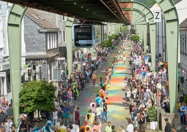 Bunte Farben auf dem Boden und eine Schwebebahn über der belebten Straße im Wuppertal Zentrum. Menschenmassen säumen die bunte Gasse.