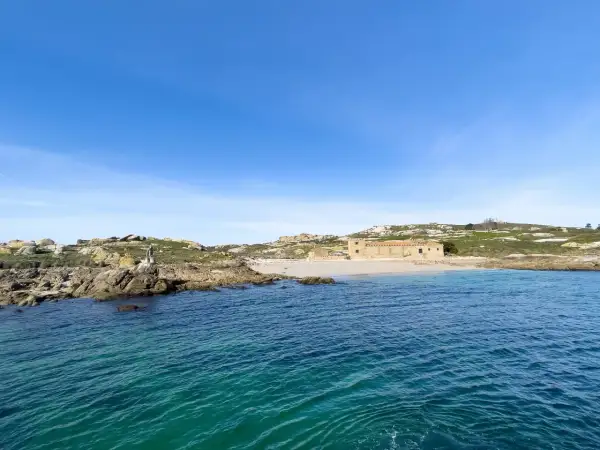 Vista de la isla de Sálvora con sus rocas, una playa de arena y un edificio de piedra bajo un cielo azul.