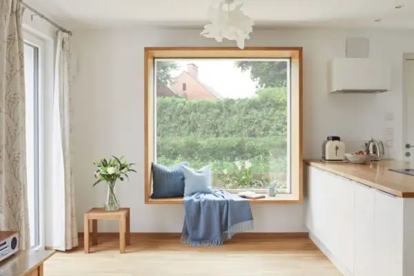 Gemütliche Fensterbank mit Kissen und Decke, ein Buch liegt offen. Das große Fenster ohne Gardinen gibt den Blick auf einen üppigen Garten frei.