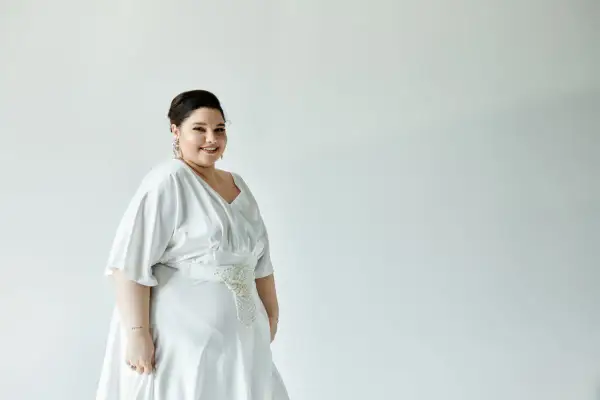 Uśmiechnięta panna młoda w pięknej, białej sukience weselnej plus size z ozdobnym pasem.