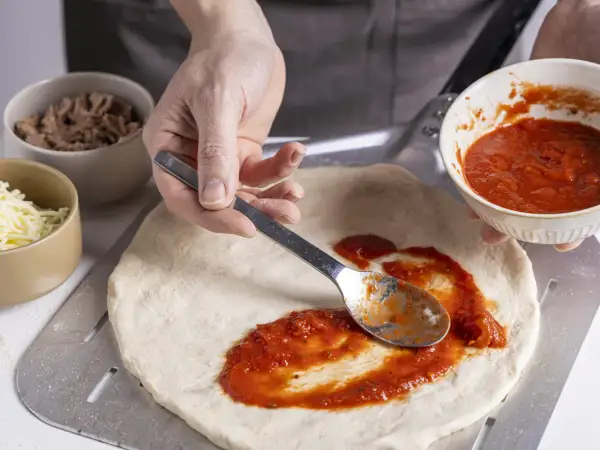 Zubereitung eines italienischen Thunfisch Pizza Rezepts: Tomatensauce wird auf den Teig gestrichen. Daneben liegen Thunfisch und Käse.
