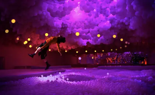 A person in a yellow shirt leaps into a ball pit at the balloon museum London, surrounded by purple clouds and yellow lights.