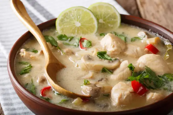 Aksamitna zupa tom kha gai z kawałkami kurczaka, grzybami i chili, udekorowana plasterkami limonki i świeżą kolendrą. Drewniana łyżka zanurzona w aromatycznym bulionie.