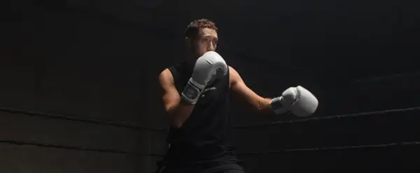 Boxer in white gloves practices a boxing slip in a dark ring, ready for the next round.